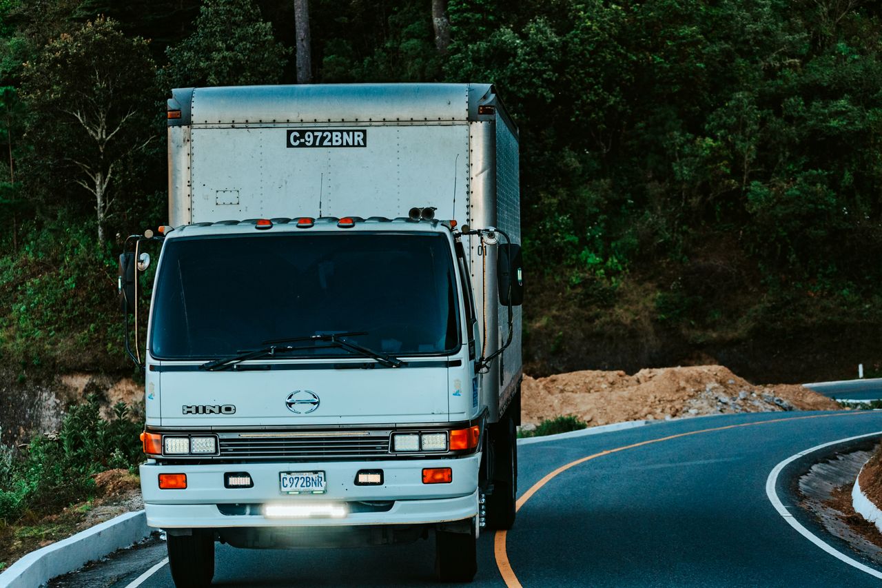 Home A white truck driving on a winding road surrounded by dense green forest plants.