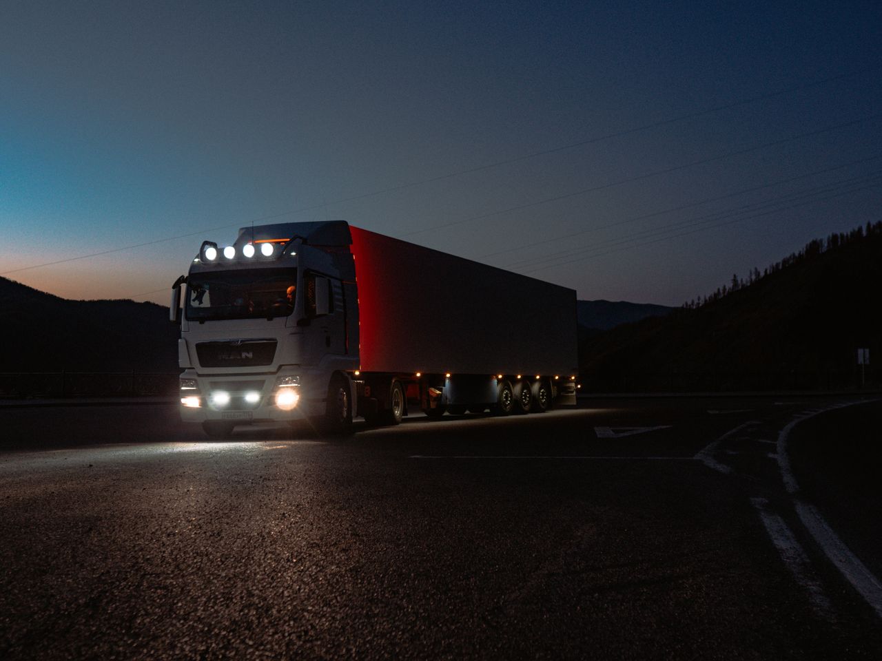 A heavy-duty truck navigates a quiet road at dusk, highlighting its illuminated path through remote mountains.